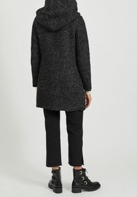 Person som bär en strukturerad mörkgrå huva kappa, svarta korta byxor och svarta snörade ankelstövlar med spännen, stående mot en enkel bakgrund.