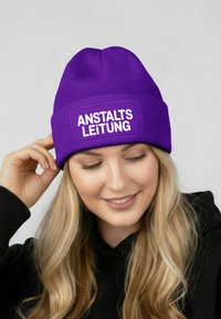 Purple knit beanie with a ribbed texture, featuring a rectangular white patch displaying the text "ANSTALTSLEITUNG."