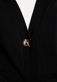 Schwarze Strickjacke mit einem auffälligen runden goldenen Verschluss als Verschluss, gefertigt aus einem glatten, strukturierten Stoff.