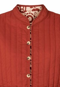 Veste matelassée rouge rouille avec encolure ronde, fermeture à boutons et doublure intérieure beige avec motif floral rouge.