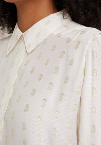Camicia bianca con colletto, caratterizzata da un motivo testurizzato di punti dorati su tutta la superficie. Il tessuto sembra leggero e leggermente trasparente.