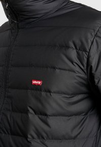 Sort oppustelig jakke med vandret quilted design, der har en høj krave og et rødt Levi's-logo på venstre side. Blød tekstur, letvægtsstof.