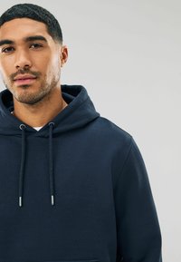 Sudadera con capucha de color azul marino, hecha de una tela suave. Presenta un bolsillo frontal tipo pouch, cordones y puños acanalados. Textura suave.