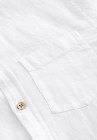Gros plan sur une chemise en lin blanche montrant une poche poitrine et un bouton beige à quatre trous sur la patte de boutonnage avant.