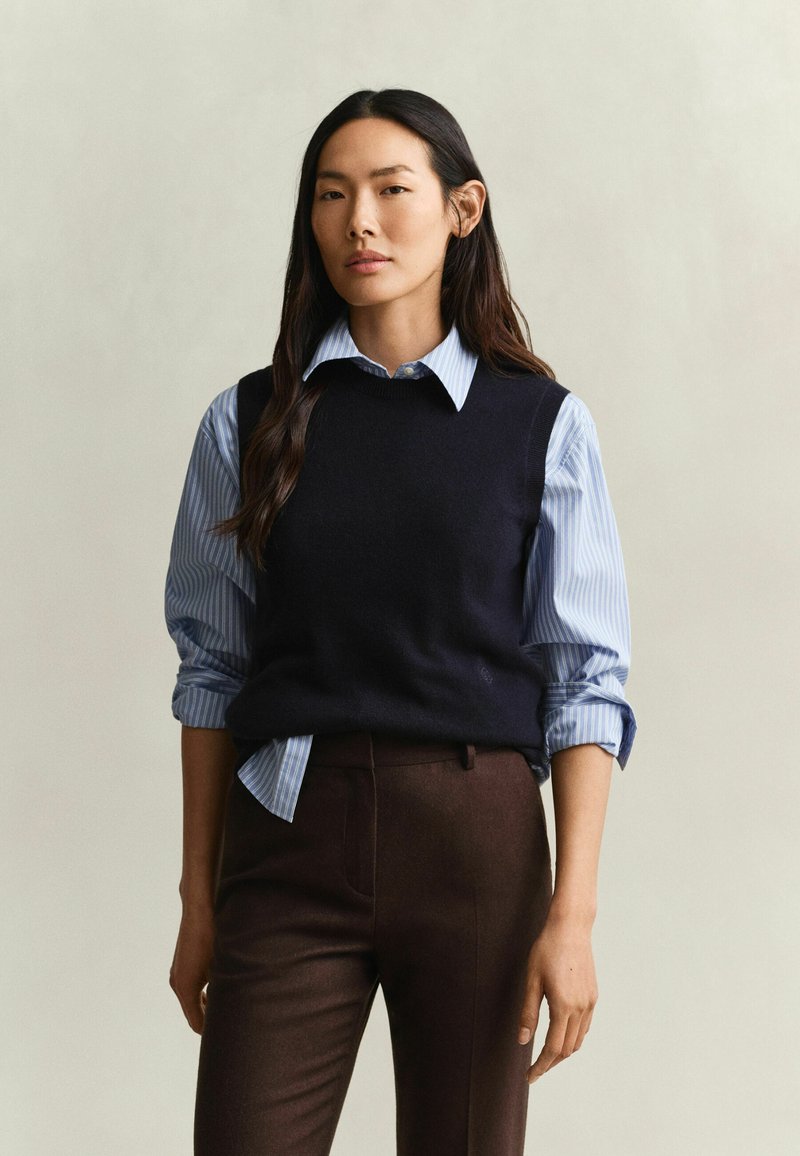 Femme aux longs cheveux foncés portant une chemise rayée bleue avec les manches retroussées, un pull sans manches noir et un pantalon marron foncé, debout devant un fond clair.