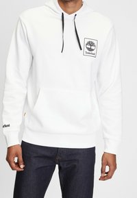 Man som bär en vit Timberland-hoodie med svarta dragsnören och logotyp på bröstet och ärmarna, ihop med mörkblå jeans.