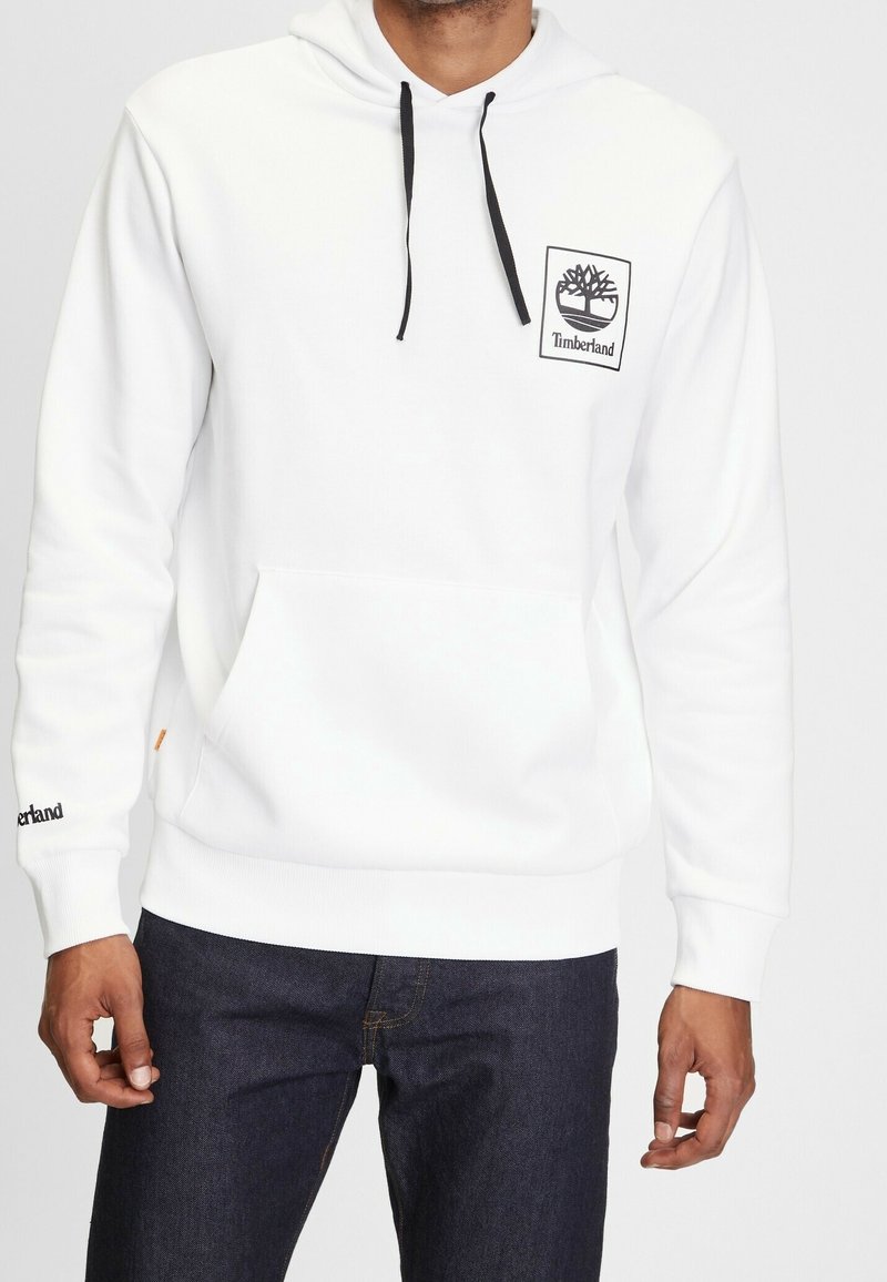 Man som bär en vit Timberland-hoodie med svarta dragsnören och logotyp på bröstet och ärmarna, ihop med mörkblå jeans.