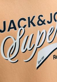 Nærbillede af beige stof med sort og hvid tekst, der lyder "JACK&JONES Superdry Siden 1990" i fede og skrå skrifttyper.