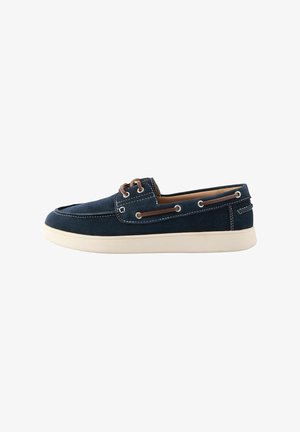 Chaussure bateau en daim bleu marine avec semelle blanche, lacets en cuir marron traversant des œillets métalliques, et détails de couture blancs.