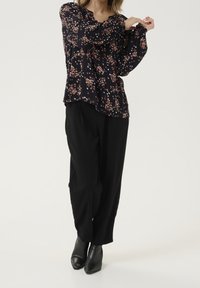 Blouse fleurie en bleu marine foncé avec des fleurs roses, jaunes et blanches ; manches longues bouffantes ; associée à un pantalon noir ample et des bottines.