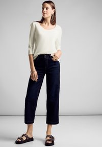 Weißes Langarmoberteil, lockere dunkle Denim-Jeans, dekorative Sandalen mit Schnallen und Ösen, dezenter Glanz auf dem Stoff, entspannte Passform.