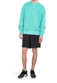 Homme portant un sweat-shirt turquoise, un short noir à genoux et des chaussures de sport noires, debout avec les bras détendus le long du corps.