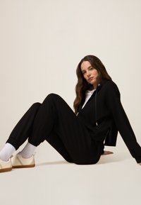 Schwarze Kapuzenjacke und passende Jogginghose aus weichem Material mit elastischen Bündchen. Verfügt über eine Fronttasche, Kordelzüge und goldfarbene Hardware-Akzente.
