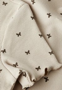 Tejido acanalado en beige con pequeños patrones de mariposas marrones. Textura suave con bordes deshilachados, que muestra un sutil detalle de diseño.