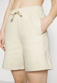 Pantalones cortos de algodón beige claro con cintura elástica, cordón, bolsillos laterales y un sutil detalle del logo en el dobladillo.