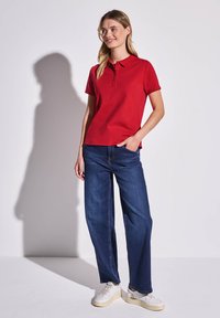 T-shirt polo rouge avec col et patte de boutonnage à trois boutons, associé à un jean droit bleu foncé et des baskets blanches. Style simple et décontracté.