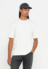 Νεαρός άνδρας με μακριές κοτσίδες, φορώντας μαύρο καπέλο, άσπρο πουκάμισο κοντομάνικο και σκούρα τζιν, κοιτάζει ελαφρώς προς το πλάι με το ένα χέρι στην τσέπη.