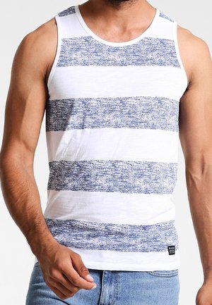 Homme portant un débardeur blanc sans manches avec de larges rayures horizontales bleu chiné et un jean bleu clair, debout avec le poing légèrement serré.