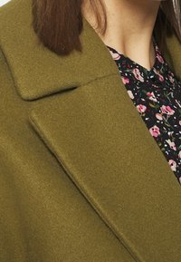 Olivgrön ullrock med breda slag, kombinerad med en blus med blomsterprint som har rosa och vita blommor på en mörk bakgrund.