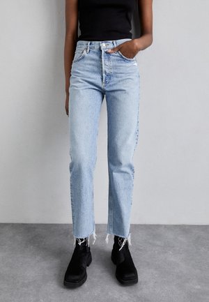 Persona con vaqueros de tiro alto de color azul claro con dobladillos deshilachados, botas negras y una camiseta sin mangas negra sobre un fondo gris liso.