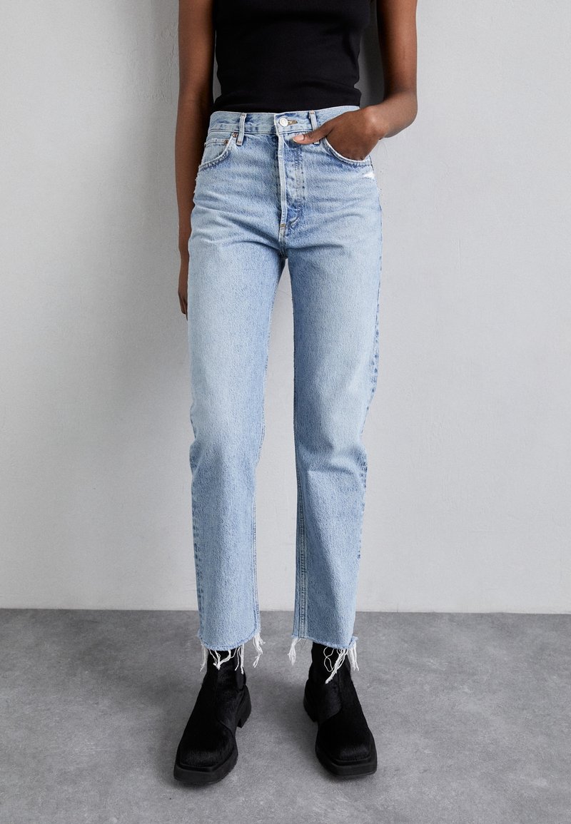 Personne portant un jean taille haute bleu clair avec des ourlets effilochés, des bottes noires et un haut noir sans manches sur un fond gris uni.