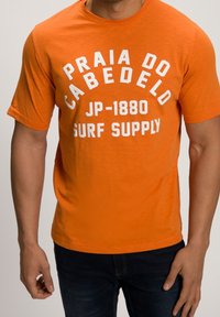 Man wearing an orange T-shirt with white text "Praia do Cabedelo JP-1880 Surf Supply" and dark blue jeans.