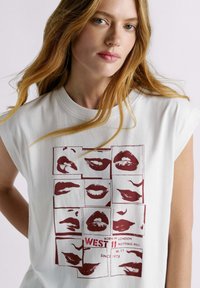 Witte katoenen T-shirt met een gedurfde afbeelding van rode lippen gerangschikt in een raster, met onderaan de tekst "WEST 11 NOTTING HILL".