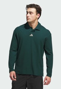 Dark green long-sleeve polo shirt with a collar, featuring a small light grey Adidas logo on the chest and a smooth fabric texture.
