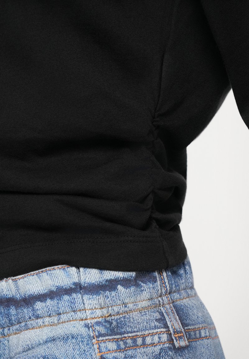 Schwarzes geripptes Oberteil mit gerafftem Seitendetail, kombiniert mit hellblauen Jeans aus Denim, die ein verwaschenes Muster und orangefarbene Nähte aufweisen.
