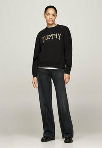 Sudadera negra con el logo "TOMMY JEANS" en dorado, combinada con jeans de mezclilla oscuros de pierna ancha y zapatos negros, de pie contra un fondo neutral.
