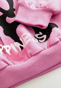 Manche et ourlet de sweat-shirt rose avec poignets côte, avec un texte graphique noir et blanc partiellement visible sur le tissu.