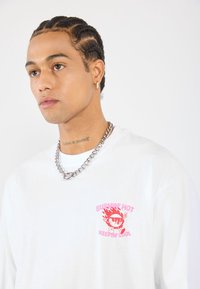 White long-sleeve t-shirt with a colorful graphic feature on the chest, a silver chain necklace, and short braided hair.