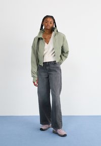 Chaqueta bomber verde claro, jeans de pierna ancha grises y zapatillas moradas. La chaqueta cuenta con un cierre frontal y un cuello alto, con una textura suave.