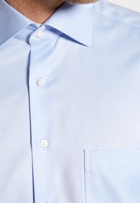 Camicia azzurro chiaro con un tessuto liscio, colletto a punta e una singola tasca sul petto. Presenta tre bottoni bianchi sulla patta.