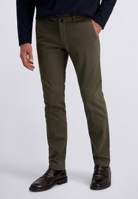 Olijfgroene chino's met een slanke pasvorm, voorzien van zijzakken en een subtiele glans, gecombineerd met donkerbruine loafers. Gladde stoftextuur.
