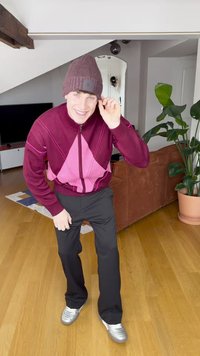 Maglione con zip color marrone scuro e rosa con motivo geometrico; pantaloni neri; sneakers grigie; berretto lavorato a maglia marrone con logo; ambiente interno con piante.