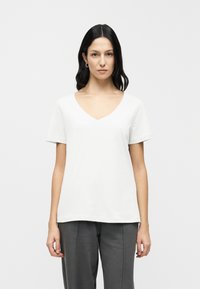 Λευκό βαμβακερό T-shirt με λαιμόκοψη V και κοντά μανίκια, με άνετη εφαρμογή. Το μοντέλο το φοράει με γκρι παντελόνι jogger.