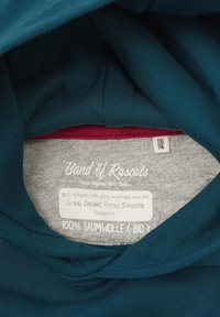 Tessuto di cotone verde-blu con etichetta grigia, contenente testo bianco sulla certificazione biologica e sulle istruzioni per la cura. Dimensione dell'etichetta 110/116.