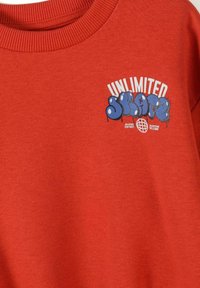 Sweat-shirt rouge avec texte en style graffiti bleu "SKATE" et texte blanc "UNLIMITED" au-dessus, plus les petites phrases "Skater District" et "Everyone Welcome".
