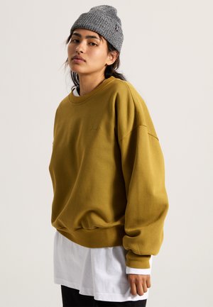 Mustard yellow sweatshirt with a crew neck, ribbed cuffs, and hem. Worn over a white long-sleeve shirt, paired with a gray knit beanie.