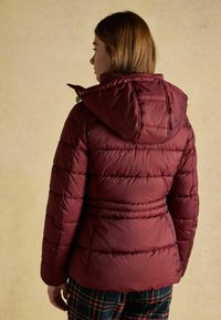 Burgundy, padded jacket with a hood, featuring horizontal stitching and a fitted waist. Model wears plaid pants, viewed from the back.