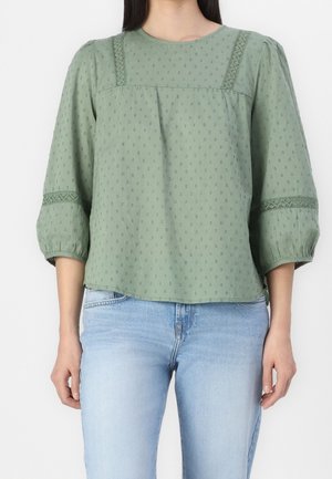 Vrouw met een lichtgroene gestructureerde blouse met opgeblazen driekwart mouwen en kantdetails, gecombineerd met lichtblauwe spijkerbroek.