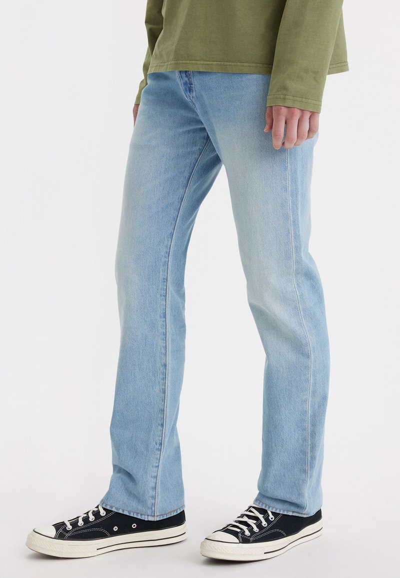 Jean en denim bleu clair avec une coupe droite, décoloration subtile et design à cinq poches, portés avec des baskets en toile noires avec des accents blancs.