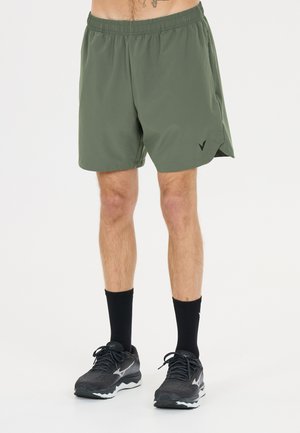 Homme portant un short de sport vert olive, des chaussettes noires et des chaussures de course noires, debout devant un fond uni.