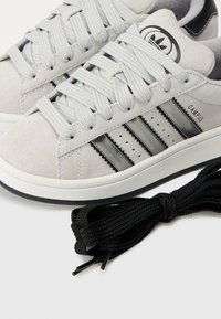 Szürke velúr tornacipők három fekete csíkkal, fehér talppal és kerek fűzőkkel. Tartalmaz egy extra fekete fűzőt. Minimalista dizájn.