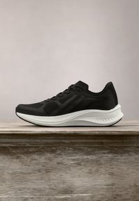 Chaussure de course athlétique noire avec tige en mesh texturé et semelle épaisse blanche coussinée reposant sur une surface en bois sur fond uni.