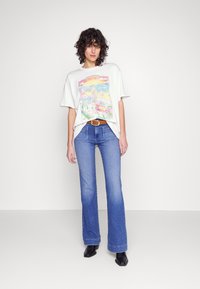 T-shirt graphique blanc avec un design scénique coloré, associé à un jean clair évasé et une ceinture marron, complété par des bottines noires.