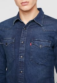Homme portant une chemise en jean bleue foncée à boutons avec poches poitrine à rabat et une petite étiquette rouge Levi's sur la poche gauche.