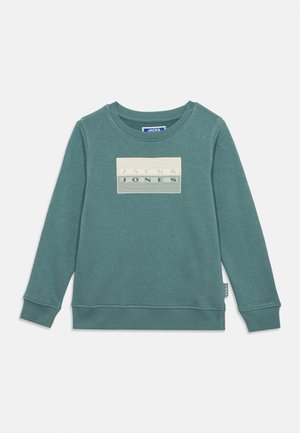 Lícny dlhý rukáv mikiny farby modrozelená s rebrovanými manžetami a lemom, s obdĺžnikovou béžovou nášivkou uprostred s nápisom "JACK & JONES".