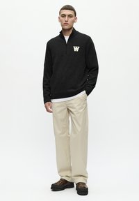 Pulover negru tricotat cu guler înalt și un logo alb "W" pe piept, asortat cu pantaloni bej și pantofi negri maronii cu șireturi.
